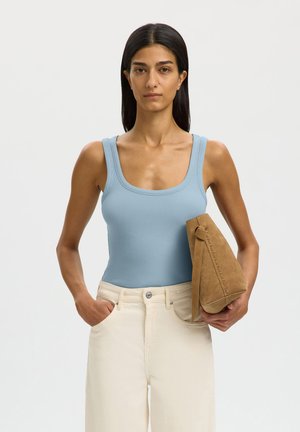 Femme aux longs cheveux foncés, portant un top sans manches bleu clair et un pantalon crème, tenant un sac à main en daim beige sur un fond uni.