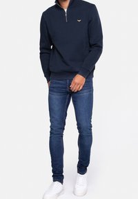 Felpa blu navy con zip e colletto alto, abbinata a jeans slim-fit blu e sneakers bianche. Presenta un logo dorato sul petto.