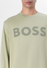 Sudadera verde claro con cuello redondo y un prominente logo "BOSS" en un tono a juego. Textura suave y corte relajado.