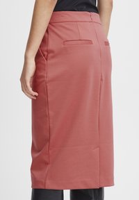 Jupe crayon de longueur intermédiaire en rose poussiéreux, fabriquée en tissu lisse, avec une couture au dos et deux poches arrière. Ceinture plate.
