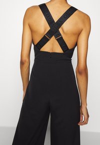 Combinaison noire avec bretelles croisées comportant un logo, une fermeture éclair à l'arrière, et un design à jambes larges pour une coupe décontractée. Texture lisse.