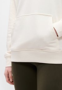 Άτομο που φοράει κρεμ φούτερ με τσέπη καγκουρό και σκούρο κολάν, με το χέρι να ακουμπά μέσα στην τσέπη.