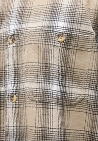 Μπεζ και καφέ πουκάμισο με καρό μοτίβο, με τσέπη στο στήθος και τρία ορατά στρογγυλά κουμπιά ραμμένα με σκούρα κλωστή.