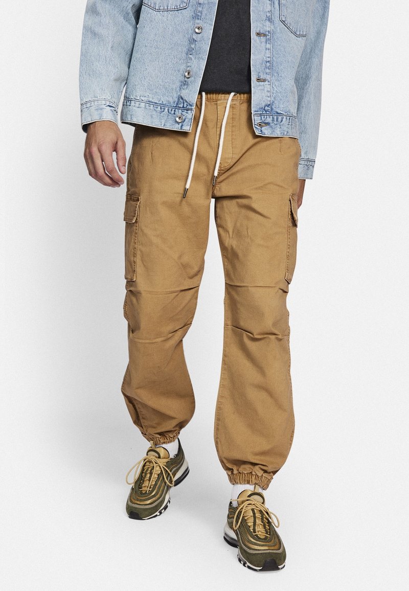 Homme portant un pantalon cargo jogger beige avec des poignets élastiques, une veste en jean claire, un t-shirt noir, des chaussettes blanches et des baskets vertes avec des lacets jaunes.