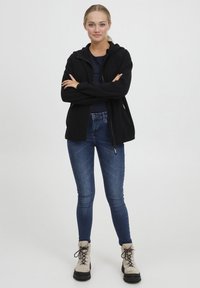 Svart fleecejakke med glidelås og hette, kombinert med mørkeblå smale jeans og beige snørestøvler med svarte såler.