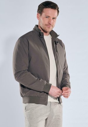 Mann trägt eine taupefarbene Bomberjacke über einem beigen Pullover und hellgrauen Hosen, schließt den Reißverschluss der Jacke vor einem schlichten hellen Hintergrund.