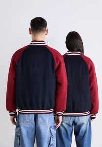 To personer står side om side med ryggen til, iført matchende navy og burgunder varsityjakker med lyseblå jeans.