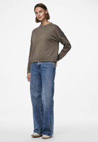 Felpa in cotone marrone con collo a giro, abbinata a jeans a gamba larga blu chiaro e scarpe beige slip-on, esposta su uno sfondo bianco.