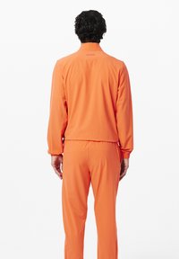 Completo sportivo arancione con giacca con zip e cintura elastica. Tessuto liscio e leggero con colletto e polsini, con un piccolo logo.
