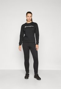 Zwarte sweatshirt met wit "Champion" logo, gecombineerd met zwarte aansluitende joggingbroeken en zwarte sportschoenen. Glad, katoenen blend stof.