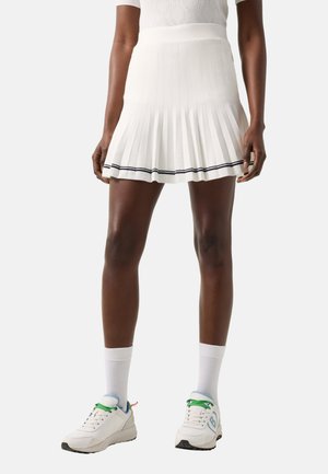 Onderlichaam van een persoon die een witte geplooide rok met zwarte zoom, witte sokken en witte sneakers met groene veters en lichtblauwe details draagt.