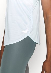 Ljusblå träningsoverdel med en texturerad yta, böjd nederkant och vit New Balance-logotyp. Bärs över grå leggings som betonar passform och rörelse.