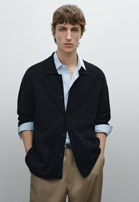 Dark navy cardigan with a collared neck, button closure, and two front pockets, worn over a light blue shirt and beige trousers.