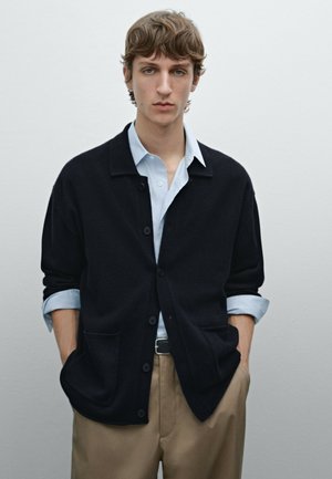 Dark navy cardigan with a collared neck, button closure, and two front pockets, worn over a light blue shirt and beige trousers.