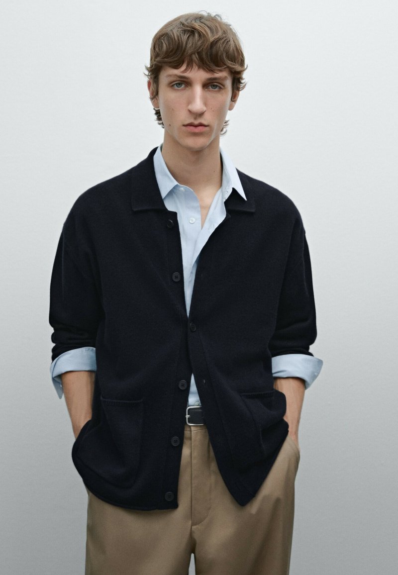 Dark navy cardigan with a collared neck, button closure, and two front pockets, worn over a light blue shirt and beige trousers.