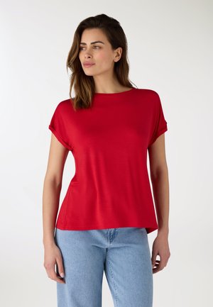 Donna con una camicia rossa a maniche corte e jeans azzurri chiari, in piedi con le mani rilassate, che guarda leggermente di lato.