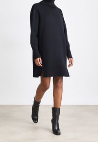 Abito maglione di colore blu navy con colletto alto e polsini a coste, dotato di spacchi laterali e vestibilità comoda, abbinato a stivaletti neri alla caviglia.