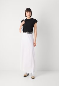 Black top with short sleeves, white flowing skirt, silver studded belt, and shiny silver and beige heeled boots. Simple bag held in hand.