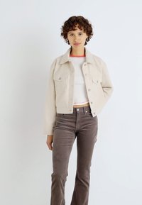 Beige cropped jakke med to frontlommer, knaplukning og en tekstureret overflade, båret over en hvid T-shirt og brune bukser.