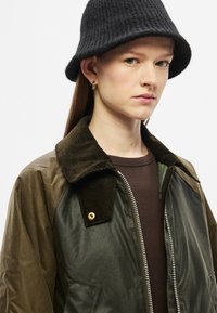 Καφέ και πράσινο μπουφάν με γιακά από κορντόρι, κλείσιμο με φερμουάρ και τσέπες. Συνδυασμένο με ένα μαύρο καπέλο τύπου bucket με υφή.