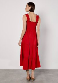 Rotes Maxikleid mit Rüschenträgern und geschmücktem Oberteil. Mit Stofflagen und einem fließenden Rock ausgestattet. Getragen mit beigen Riemchenheels.