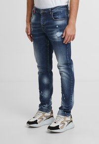 Modré oprané džíny s úzkým střihem, s vybledlými místy a menšími trhlinami. Vzájemně kombinované s bílými a béžovými teniskami s černými akcenty.