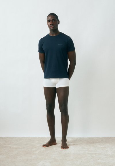 Νεαρός άνδρας όρθιος, ξυπόλητος σε χαλί, φοράει μπλε σκούρο μπλουζάκι και λευκά βαμβακερά εσώρουχα μπροστά σε λευκό απλό τοίχο.