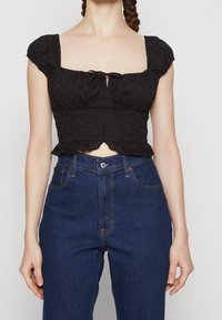 Černý bavlněný crop top s krátkými nafouklými rukávy, čtvercovým výstřihem, předním ozdobným vázáním a vlnkovaným spodním lemem. V kombinaci s modrými džíny s vysokým pasem.