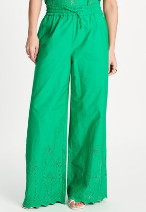 Vrouw die felgroene, wijde broek draagt met elastische tailleband en trekkoord, met een geschulpte zoom en uitgesneden palmboom patronen.