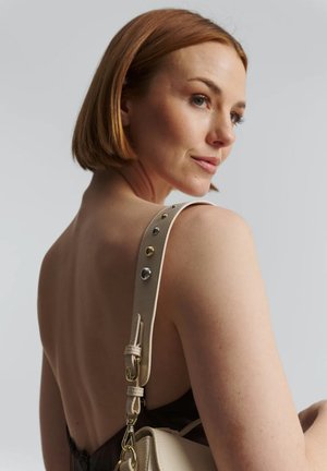 Beige leren handtas met gouden en zilveren hardware, met een gestructureerd ontwerp, verstelbare band en een textuurafwerking.