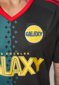 Camiseta de fútbol negra con acentos rojos, que presenta un diseño de cuello en V. El frente tiene un patrón azul verdoso, un logo amarillo "GALAXY" y el texto "LOS ÁNGELES".