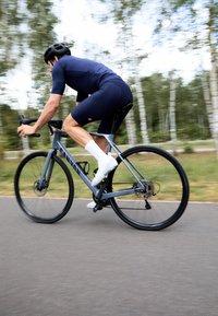 Bici da corsa grigia in movimento con ruote nere. Ciclista in maglia blu e pantaloni neri, con casco nero e calze bianche. Alberi sullo sfondo.
