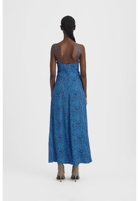 Robe maxi bleue à motif, bretelles fines et coupe ample, associée à des bottes à talons hauts beige clair. Texture de tissu lisse.