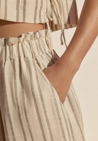 Buste de femme avec la main dans la poche portant un pantalon en lin couleur crème et un top court assorti avec fines rayures verticales marron.