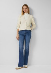 Cremefarbener, gerippter Cardigan mit Knöpfen, getragen über einem weißen Rollkragenpullover; kombiniert mit blauen Schlagjeans und hellen Sneakers.