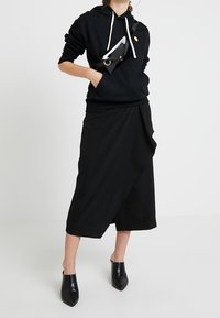 Svart huvtröja med vita dragsnören, kombinerad med en svart asymmetrisk kjol. Accessoarer inkluderar en liten svart väska som bärs diagonalt. Svarta ankelskor.