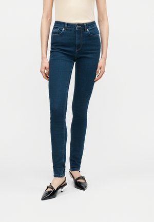 Žena oblečená v tmavomodrých, vysokým pásom priliehavých džínsoch a čiernych lodičkách s otvorenou pätou a špičatou špičkou, stojaca pred jednoduchým svetlým pozadím.