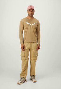 Beige Langarmhemd mit weißen Akzenten, tansfarbene Cargo-Hosen und eine pinke Mütze, kombiniert mit grauen Wanderschuhen.