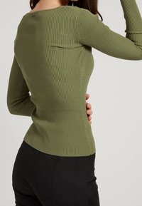 Ribbed, long-sleeve top in olive green, featuring a boat neck and fitted design. Smooth texture with a slightly curved hem.