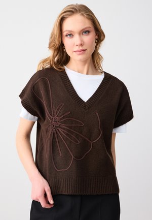 Kobieta o blond włosach ubrana w ciemnobrązową dzianinową kamizelkę z dużym kwiatowym haftem na białej koszulce z krótkim rękawem na jednolitym tle.