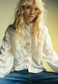 Camicia bianca a maniche lunghe ricamata con motivi floreali; collane d'oro a strati; modello che indossa jeans blu in posa seduta.