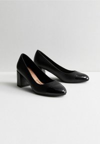 Zapatos de tacón de cuero negro con punteras de cuero patentado, tacones cuadrados, textura suave y un diseño sencillo.