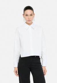 Camisetta bianca corta con un colletto strutturato decorato con strass; maniche lunghe con bottoni decorativi sui polsini. Abbinata a jeans neri.
