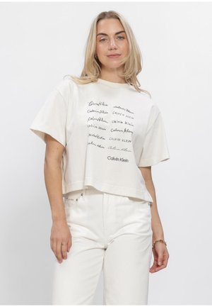 Vrouw draagt een wit Calvin Klein cropped T-shirt met meerdere logostijlen en witte high-waisted jeans tegen een effen achtergrond.