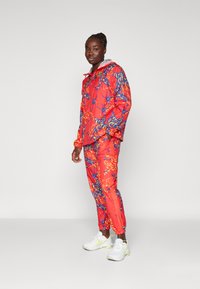 Chaqueta de lluvia roja y pantalones a juego con un patrón floral azul y amarillo, puños elásticos y capucha. Un par de zapatillas blancas con acentos verdes.