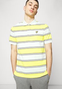 Stripet polo skjorte med gule, hvite og svarte striper, kravehals og en liten logo på venstre bryst. Glatt bomullstoff.