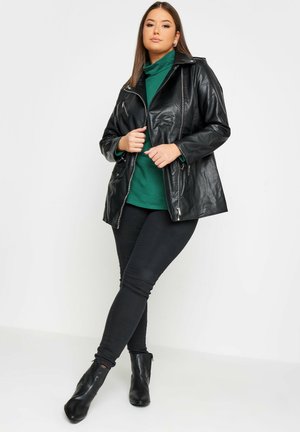 Zwarte leren jas met een ritssluiting aan de voorkant, zijzakken en een gestructureerde kraag, gedragen over een groene coltrui en zwarte skinny jeans.