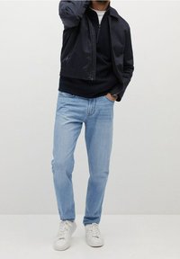 Veste légère marine portée sur un pull sombre, associée à un jean bleu clair et des baskets blanches. Coupe classique avec un style décontracté.