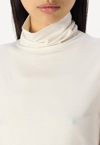 Kremfarget høyhalset topp laget av glatt stoff, med en tett, brettet krage og en tettsittende design. Myk tekstur med minimale sømmer.