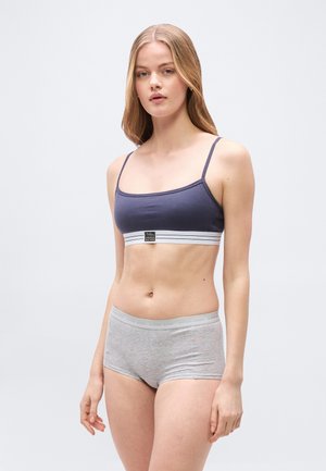 Νεαρή γυναίκα με μακριά ξανθά μαλλιά, φοράει μπλε αθλητικό σουτιέν με λευκή ελαστική ταινία και ανοιχτό γκρι εφαρμοστά σορτς, στέκεται σε ουδέτερο φόντο.